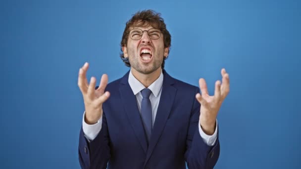 Frustrated young man, decked out in a business suit, furiously shouting his heart out. crazy mad expression with arms flying high, set against a stark isolated blue background. - Footage, Video