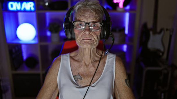 Vážná šedovlasá starší žena fáborka zabraná do noční digitální hry doma, uvolněná, ale soustředěná, sedící se sluchátky a gamepadem v herně - Fotografie, Obrázek