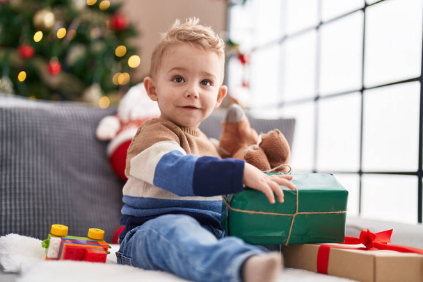 Очаровательный малыш распаковывает рождественский подарок сидя дома на диване - Фото, изображение