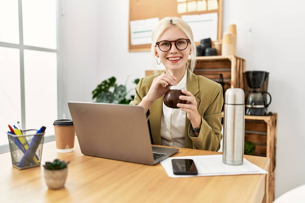 Молодая блондинка бизнес-работница, использующая ноутбук питьевой мат вливания в офисе - Фото, изображение