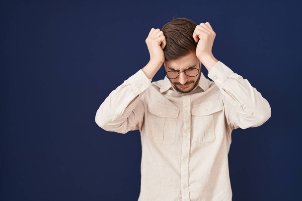 Hispánec s vousy stojí nad modrým pozadím trpí bolestí hlavy zoufalý a vystresovaný, protože bolest a migréna. Ruce za hlavu.  - Fotografie, Obrázek