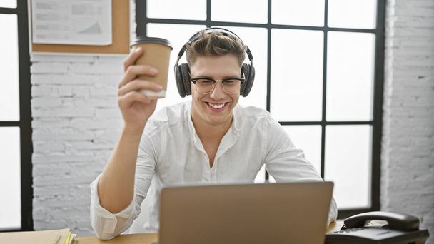 若い慎重な男ビジネスワーカー オフィスでコーヒーを保持するラップトップとヘッドフォンを使用して - 写真・画像