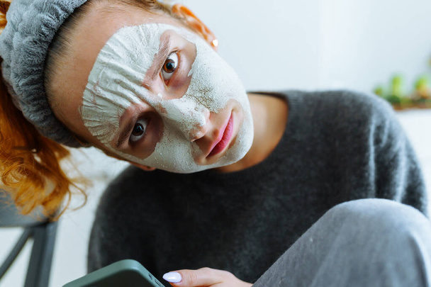 retrato cara joven mujer con máscara cosmética, con banda de pelo, mirando a la cámara, sosteniendo el teléfono móvil - Foto, imagen