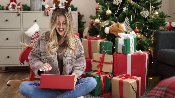 jong blond vrouw uitpakken cadeau zitten door christmas boom thuis - Foto, afbeelding