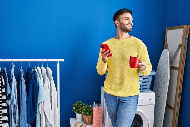 洗濯室で洗濯機を待っているスマートフォンでコーヒーを飲んでいる若いヒスパニック男性 - 写真・画像