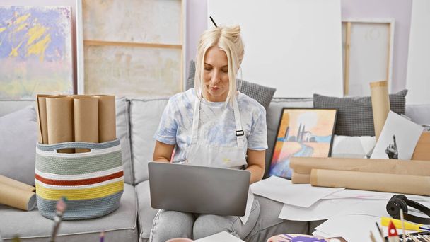 Serieuze jonge blonde kunstenaar, geconcentreerd en gefocust, zittend op studio bank met laptop voor online kunst klasse, ondergedompeld in de wereld van tekenen en schilderen. - Foto, afbeelding