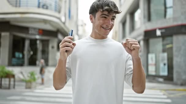 Joven adolescente hispano, celebrando exuberantemente su victoria callejera en su teléfono inteligente, mientras pasea por la ciudad. chico conocedor de la tecnología, la felicidad irradiando, participar en una conversación en línea emocionante. - Imágenes, Vídeo