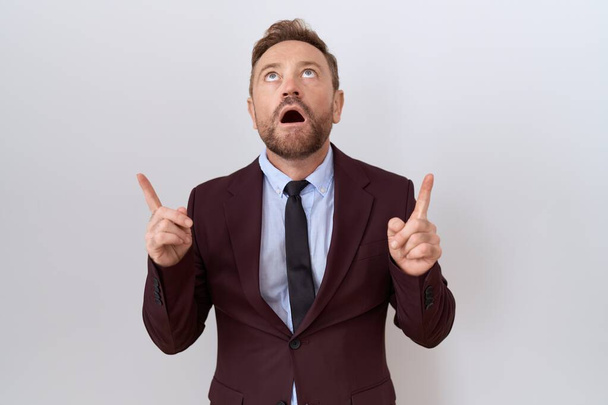 Middle age business man with beard wearing suit and tie amazed and surprised looking up and pointing with fingers and raised arms.  - Photo, Image