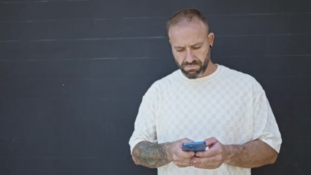Fröhlicher junger Mann, der in ein digitales Gespräch vertieft ist, SMS auf seinem Handy verschickt und glücklich über einen isolierten schwarzen Hintergrund lächelt - Filmmaterial, Video
