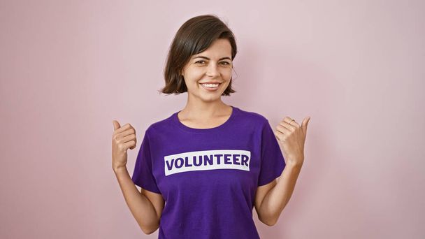 Enthusiastische junge hispanische Freiwillige mit selbstbewusster Daumen-hoch-Geste, kurzen Haaren und Charity-Uniform, die vor rosa Hintergrund isoliert von Ohr zu Ohr grinst - Foto, Bild