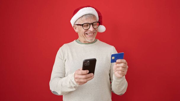 Середньовіччя сіро-волохатий чоловік робить різдвяні онлайн-покупки зі смартфоном над ізольованим червоним тлом - Фото, зображення