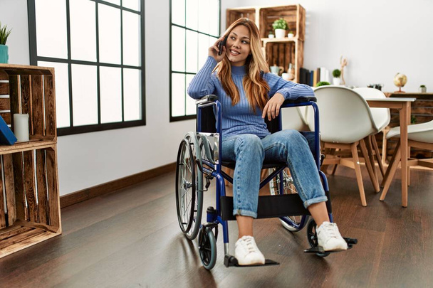 Young caucasian woman talking on smartphone sitting on wheelchair at home - Photo, Image