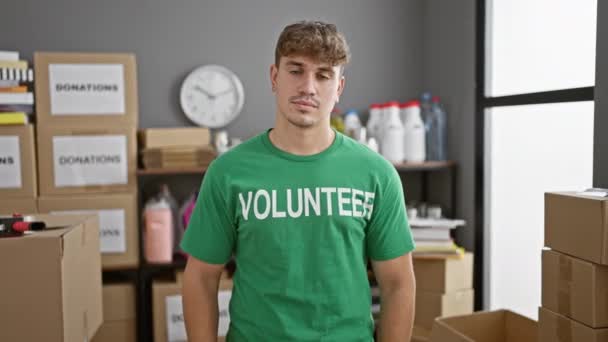 Glimlachende jonge Spaanse man die vol vertrouwen vrijwilligerswerk doet bij een liefdadigheidscentrum, duimen opgeeft ter ondersteuning, groot staat in uniform, midden in donatiedozen. - Video