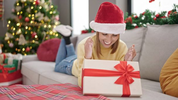 Junge blonde Frau packt Geschenk aus, das zu Hause auf Sofa am Weihnachtsbaum liegt - Foto, Bild