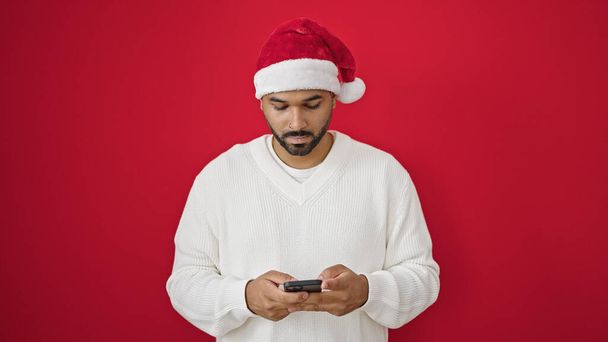 Afrikaanse amerikaanse man het dragen van kerst hoed met behulp van smartphone over geïsoleerde rode achtergrond - Foto, afbeelding