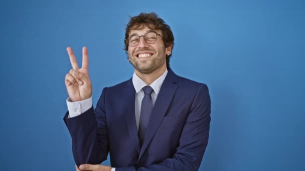 Winnende glimlach! Jonge man in zakenpak knipoogt, knippert met een vriendelijk vredesbord, zijn twee vingers vormen een 'v' voor de overwinning. geïsoleerd op blauwe achtergrond. - Video