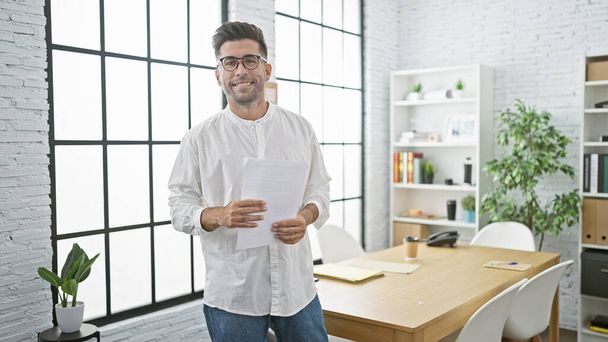 La joie rayonne d'un jeune homme hispanique à succès au travail, tenant avec confiance des documents d'affaires, souriant dans le fond du bureau. - Photo, image