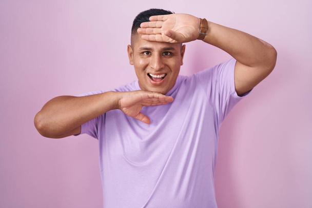 Joven hombre hispano de pie sobre fondo rosa sonriente alegre jugando a echar un vistazo a un boo con las manos mostrando la cara. sorprendido y salido  - Foto, Imagen
