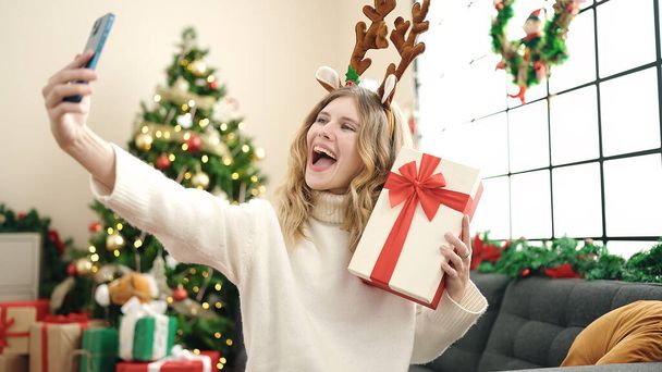 Νεαρή ξανθιά γυναίκα κάνει selfie από το smartphone κρατώντας χριστουγεννιάτικο δώρο στο σπίτι - Φωτογραφία, εικόνα