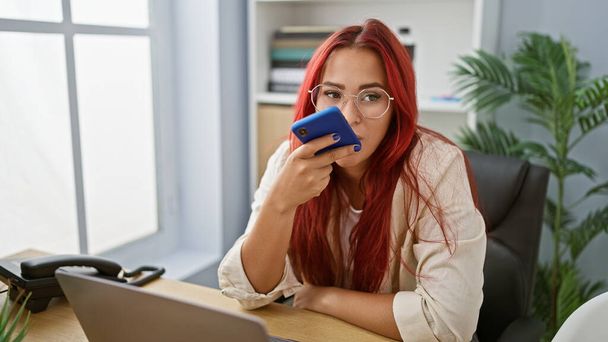 Élénk fiatal vörös hajú dolgozó nő, diadalmasan hangüzenetet küld okostelefonról az irodában - Fotó, kép