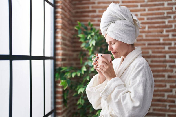 Young blonde woman wearing bathrobe smelling cup of coffee at bedroom - Photo, Image