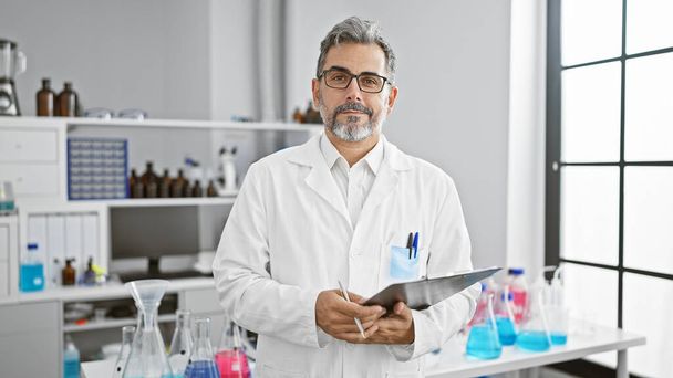 Schöner, grauhaariger junger hispanischer Mann, der sich auf die medizinische Forschung konzentriert und drinnen im Wissenschaftslabor mit Klemmbrett steht - Foto, Bild