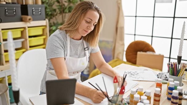 Λαμπερή, νεαρή ξανθιά καλλιτέχνιδα ζωγραφίζει χαρούμενα στο touchpad της, ρίχνοντας την ψυχή της στον ψηφιακό καμβά στο άνετο γραφείο της στο στούντιο τέχνης - Φωτογραφία, εικόνα