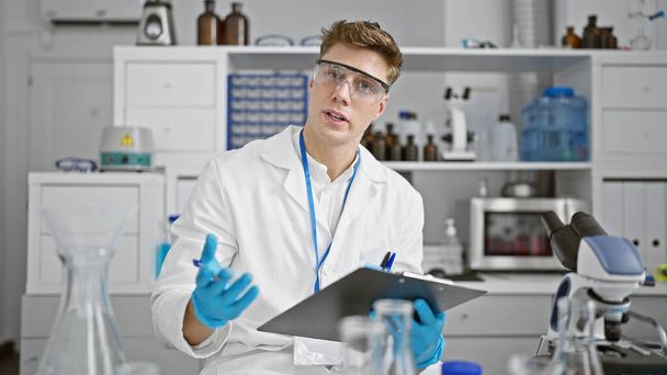 Atractivo y sonriente joven científico caucásico, sujetando el portapapeles y hablando apasionadamente de su innovadora investigación en el bullicioso laboratorio - Foto, Imagen