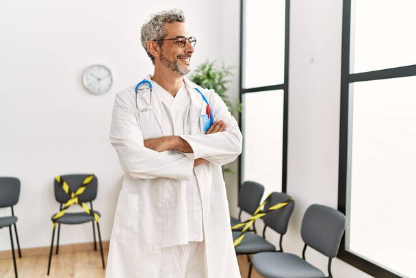 Homme moyen aux cheveux gris médecin souriant confiant debout avec les bras croisés geste dans la salle d'attente de la clinique - Photo, image