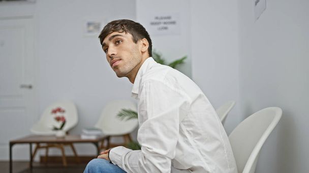Joven hispano de cara seria, mirando a su alrededor atentamente mientras está sentado en una sala de espera, exudando profesionalismo. - Foto, imagen