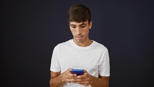 Incrível jovem hispânico surpreende com um gesto ok, mensagens de texto alegremente em seu smartphone, isolado em um fundo preto - Filmagem, Vídeo