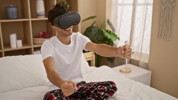Aantrekkelijke jonge Spaanse man, gamer guy met behulp van vr bril, vol vertrouwen genieten van futuristische cyber driving game als hij zit op bed, indoor home setting - Video