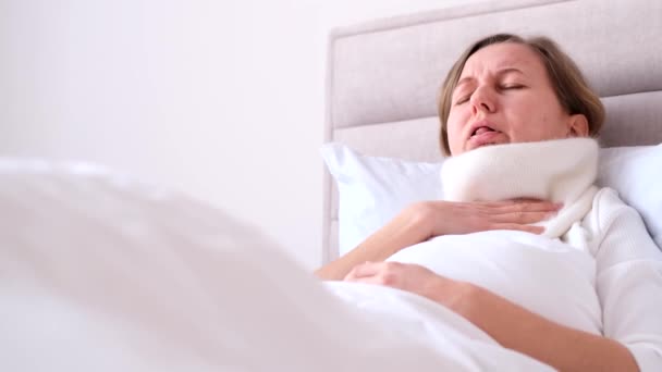 Een zieke vrouw ligt in bed in een dikke sjaal en heeft een sterke hoest. Een zieke vrouw raakt haar nek aan met haar hand, heeft keelpijn. - Video