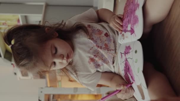 Ένα κοριτσάκι ζωγραφίζει με ένα ροζ μαρκαδόρο καθισμένο στον καναπέ. Παιδική ηλικία, δημιουργικότητα και χόμπι έννοια. - Πλάνα, βίντεο