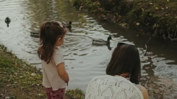 Mother with her little daughter observing ducks on lake in an autumn park outdoors. Family walk in nature, motherhood - Footage, Video