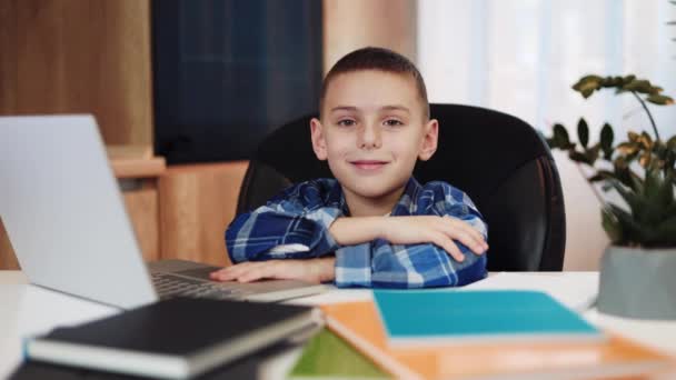 Portrait of positive school boy dressed in checked shirt keeping hands crossed on table while sitting in comfy chair. Adorable caucasian guy smiling and looking at camera with laptop and books around. - Footage, Video