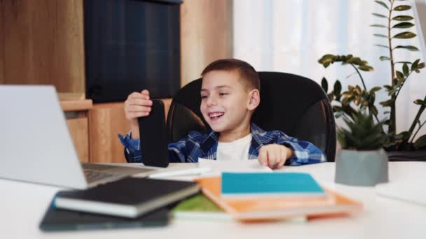 Biały chłopiec siedzi przy biurku z przenośnym komputerem i książkami trzymając komórkę w ręku. Dziecko korzystające z urządzenia cyfrowego do różnych działań online jako e-learning lub rozrywka ay cozy home. - Materiał filmowy, wideo