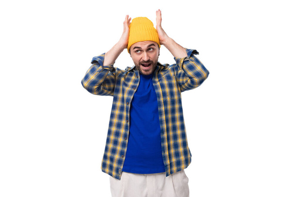 νεαρός μοντέρνος μελαχρινός άντρας με άγρια γενειάδα και μουστάκι σε καπέλο και πουκάμισο κρατώντας το κεφάλι του σε λευκό φόντο με χώρο για φωτοτυπίες. - Φωτογραφία, εικόνα