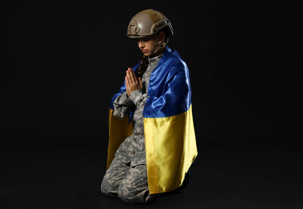 Joven soldado de uniforme con bandera de Ucrania rezando sobre fondo negro - Foto, Imagen