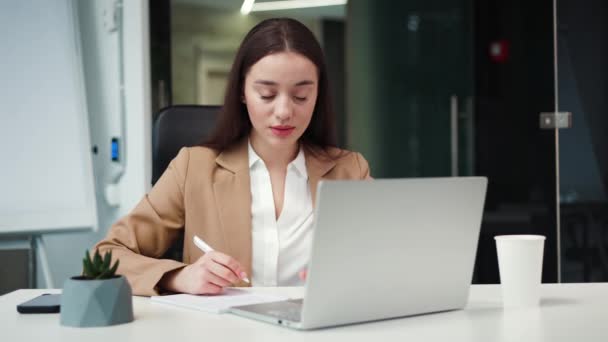 Attractive dark haired lady wearing stylish suit sitting near wireless laptop in contemporary office. Successful caucasian manager filling out application and solving working details in cozy cabinet. - Footage, Video