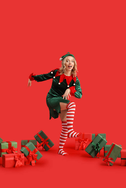 Vtipná mladá žena oblečená jako elf s vánoční dárky na červeném pozadí - Fotografie, Obrázek