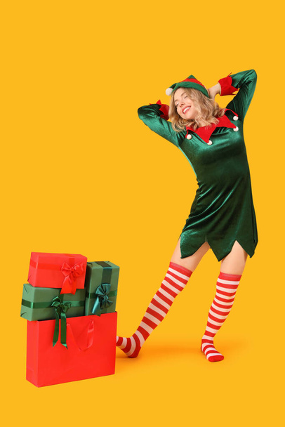 Belle jeune femme habillée en elfe avec des cadeaux de Noël s'étendant sur fond jaune - Photo, image