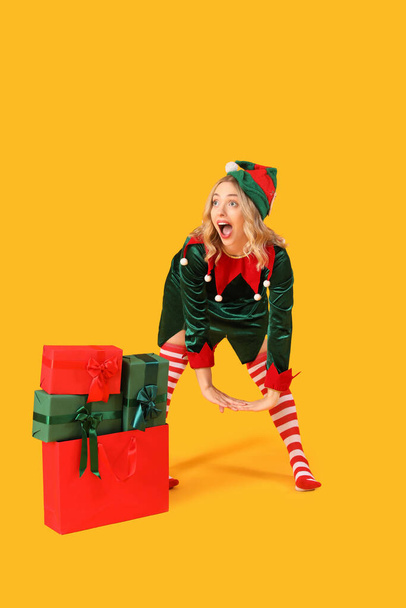 Schockierte junge Frau als Elfe verkleidet mit Weihnachtsgeschenken auf gelbem Hintergrund - Foto, Bild