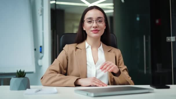 Gözlüklü ve takım elbiseli iyimser kadın girişimcinin portresi modern ofiste el ele tutuşurken kablosuz dizüstü bilgisayarın yanında oturuyor. Başarılı kariyer ve teknoloji kavramı. - Video, Çekim