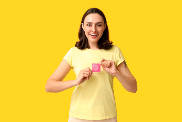 黄色の背景にコンドームを持つ美しい若い女性. 安全なセックスコンセプト - 写真・画像