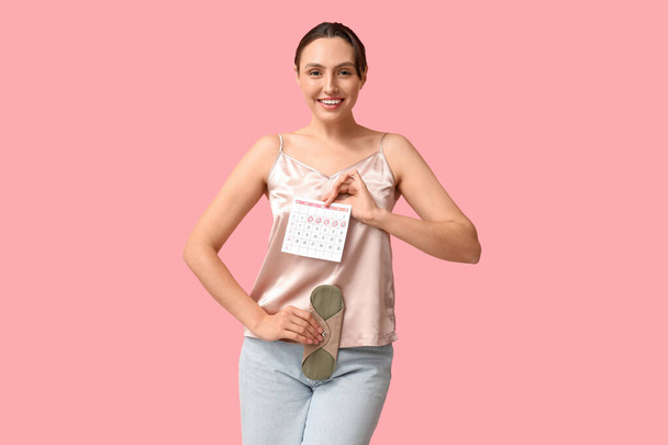 Piękna młoda kobieta trzyma podkładkę wielokrotnego użytku i kalendarz z zaznaczonymi dniami miesiączki na różowym tle - Zdjęcie, obraz