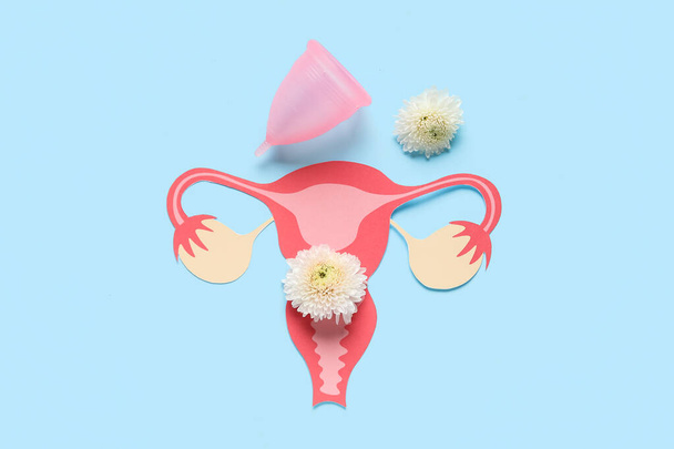 Menstruatiecup met papieren baarmoeder en bloemen op blauwe achtergrond - Foto, afbeelding