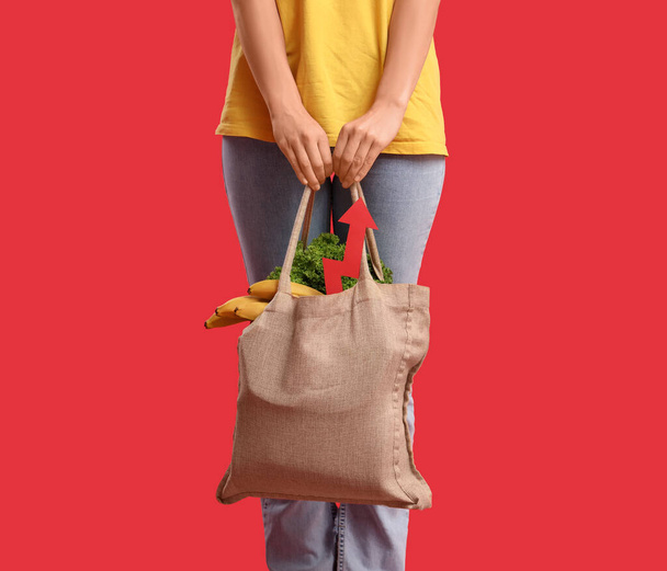 Νεαρή γυναίκα με σακούλα με φαγητό και βέλος στο κόκκινο φόντο. Έννοια της αύξησης τιμών - Φωτογραφία, εικόνα