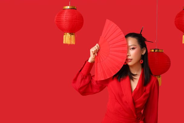 Mooie jonge Aziatische vrouw met lantaarns en waaier op rode achtergrond. Chinees nieuwjaarsfeest - Foto, afbeelding