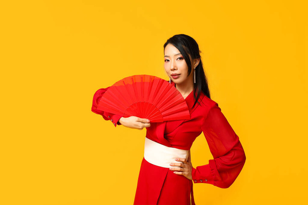 Beautiful young Asian woman with fan on yellow background. Chinese New Year celebration - Photo, Image